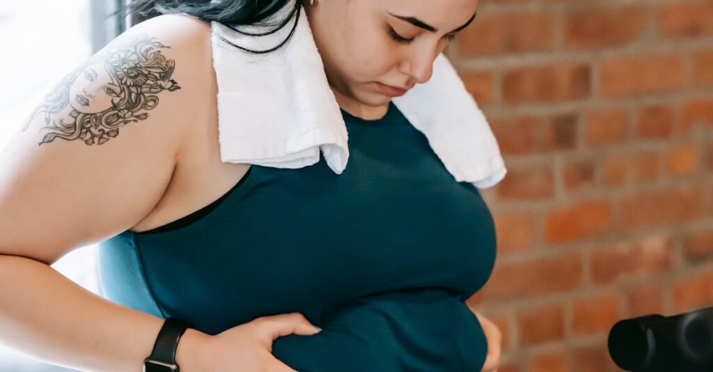 Upset young obese female in sportswear leaning on windowsill and desperately looking at stomach